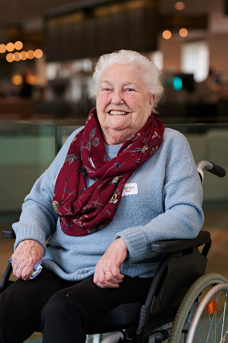 Een ouderen vrouw met blauwe trui en paarse sjaal zit in een rolstoel. Ze kijkt lachend richting de camera.