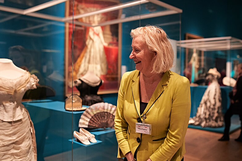 Een gastvrouw met geel colbert staat naast een vitrine en kijkt met een glimlach naar kunstwerken