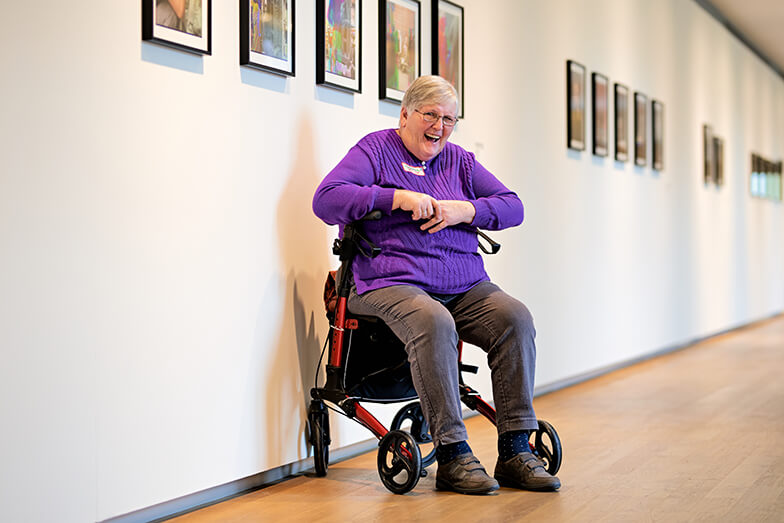 Een oudere vrouw met paarse trui zit lachend op haar rollator. Achter haar hangen verschillende foto's aan de muur.