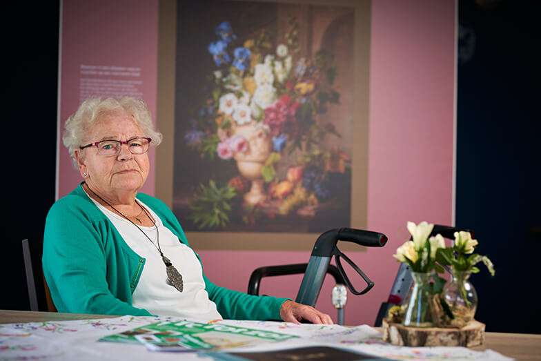 Een bewoner zit op een stoel. Naast haar staat haar rollator. Ze zit voor een replica met daarop een grote bos kleurige bloemen.