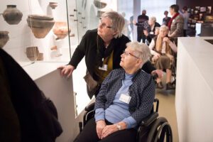 Een oudere vrouw in rolstoel kijkt samen met haar begeleider naar een vitrine vol kunstschatten.